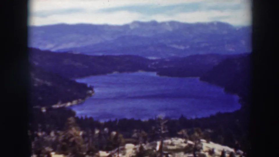 1937 Lake Tahoe home-movie frame showing blue water, dark wooded slopes, and distant mountain ridges under a pale sky.