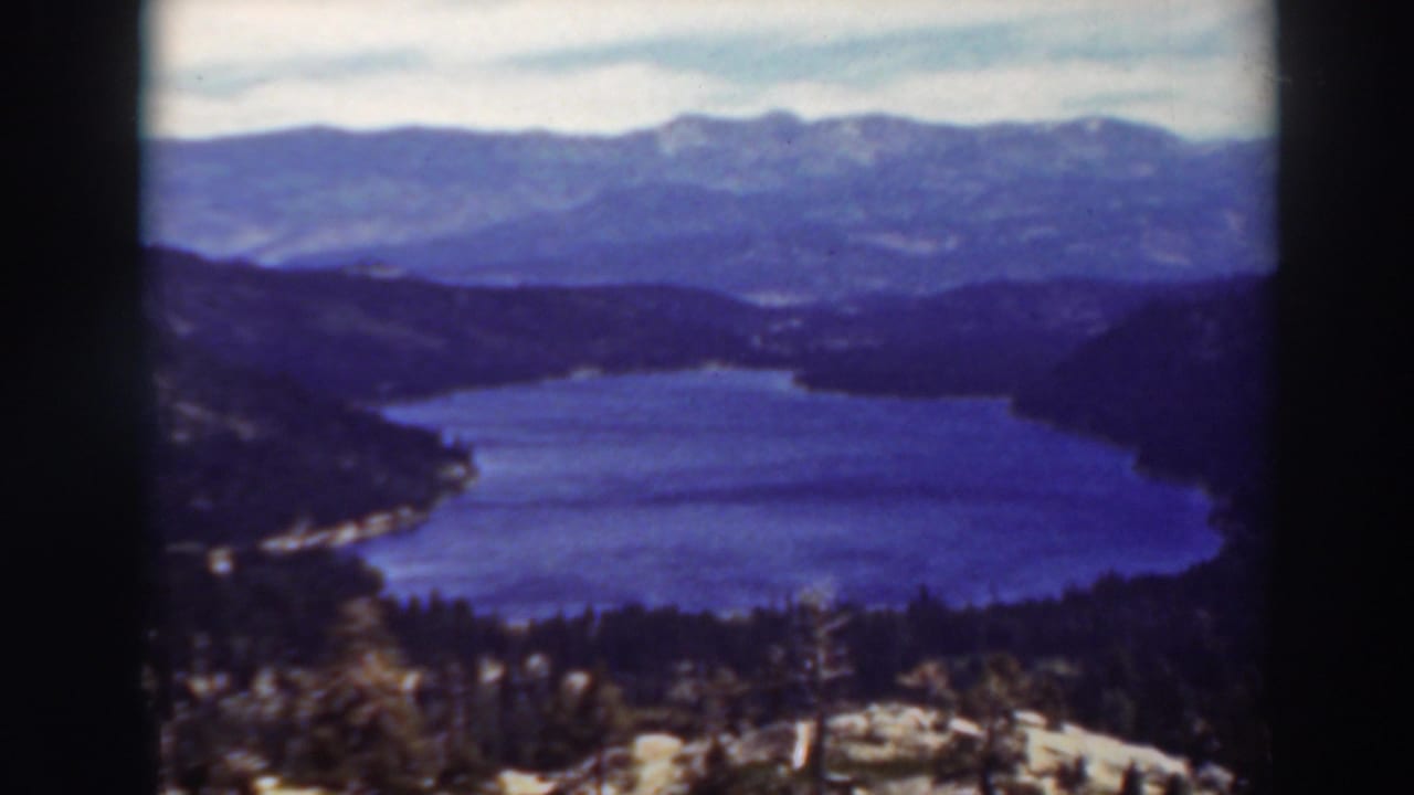 Blue lake surrounded by wooded hills and distant mountains under a cloudy sky.