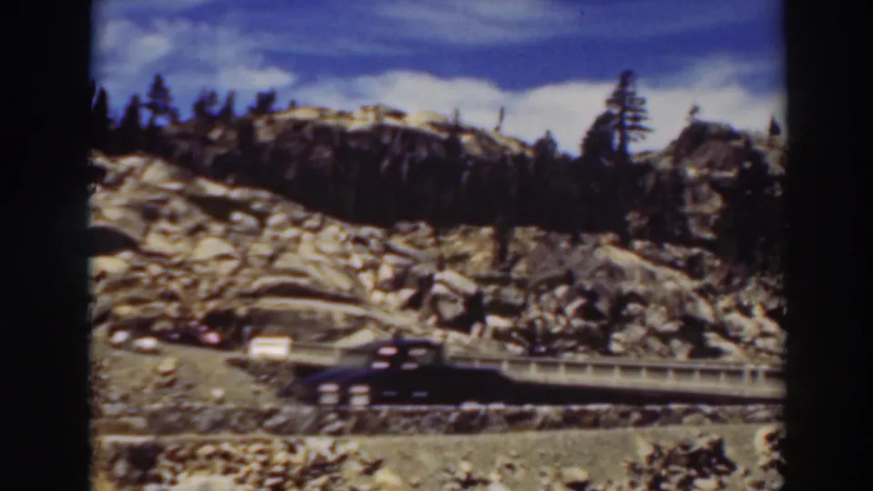 1937 Lake Tahoe road scene with a dark car moving past a small bridge beneath rocky hills and pine trees.