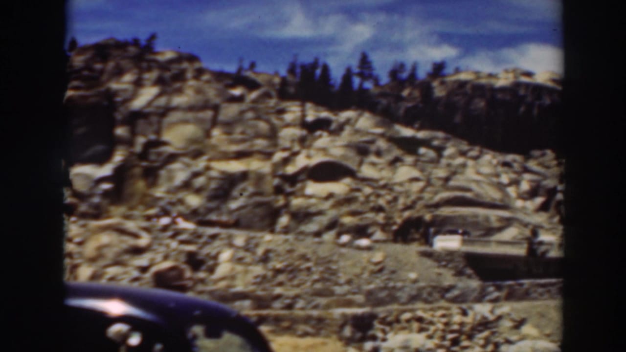Dark car roof passes below a rocky hillside with pine trees and road traffic.
