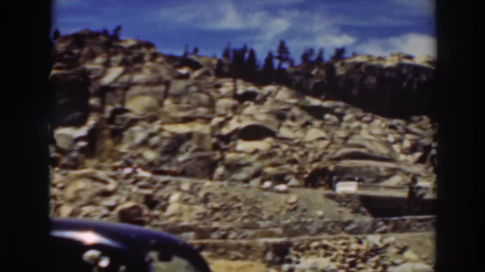 1937 Lake Tahoe home-movie frame showing a dark sedan foreground, rocky hillside, pine trees, and cars on a mountain road.
