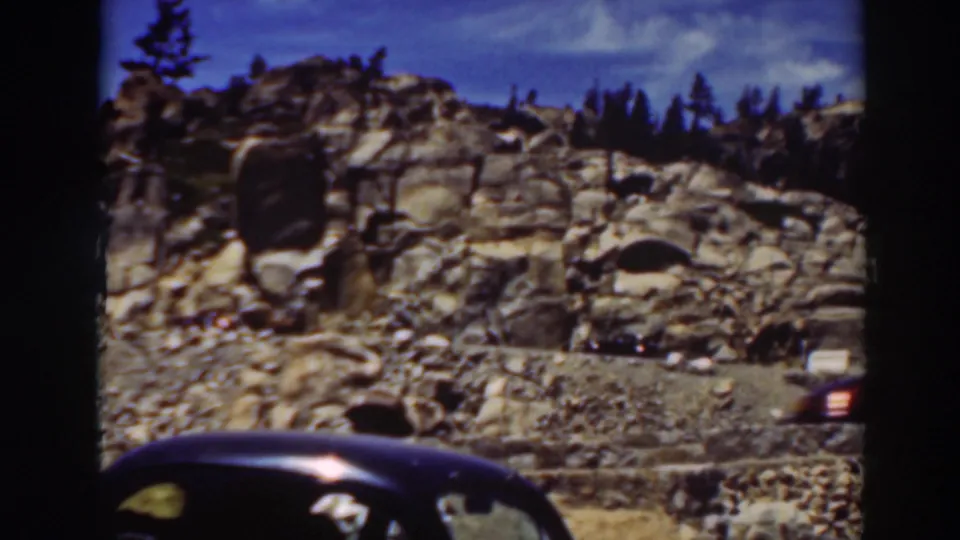 Lake Tahoe mountain road scene with rocky terrain, pines, and partial views of period cars in the foreground.