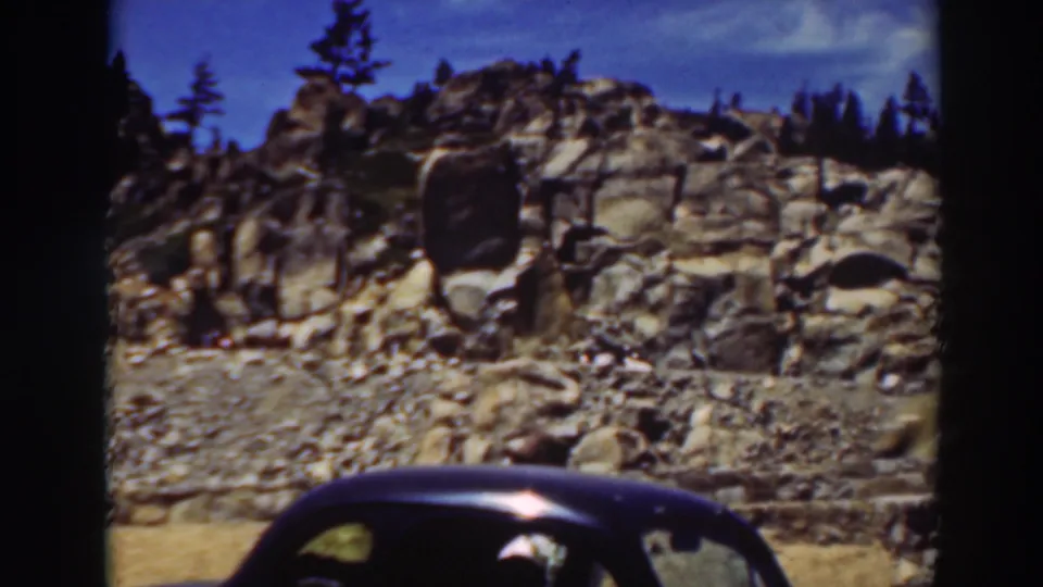1937 Lake Tahoe frame with a boulder-strewn slope, pine trees, and a dark sedan moving across the foreground.