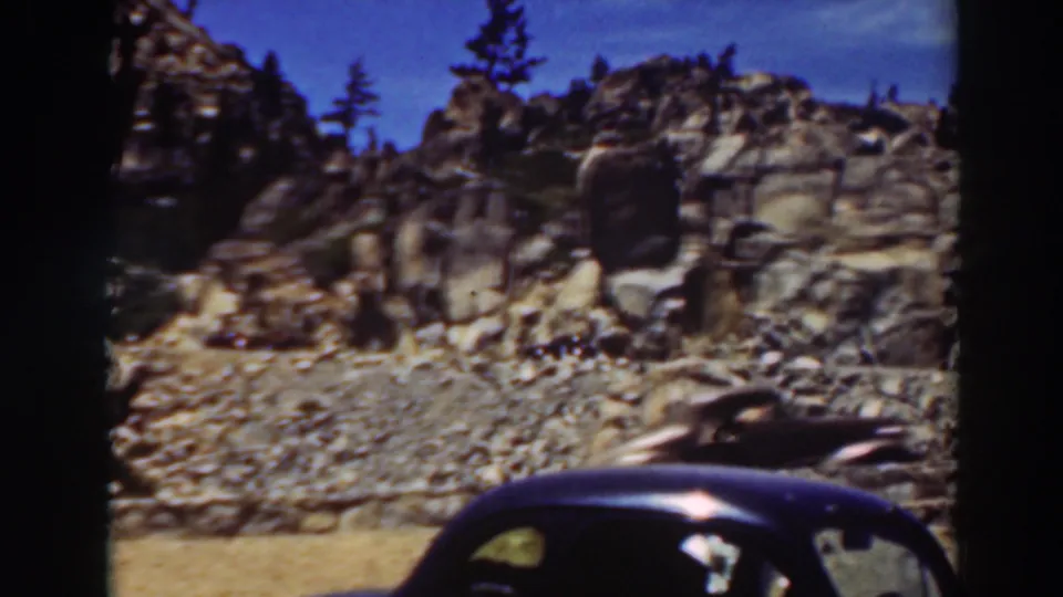 Lake Tahoe 8mm home-movie frame showing passing cars beneath a rugged rock face and pine-covered ridge.