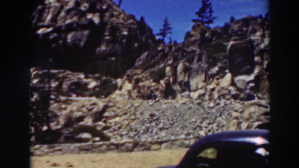 1937 Lake Tahoe travel footage frame with sunlit rock formations, scattered pine trees, and a dark vintage automobile partly entering the lower right foreground.