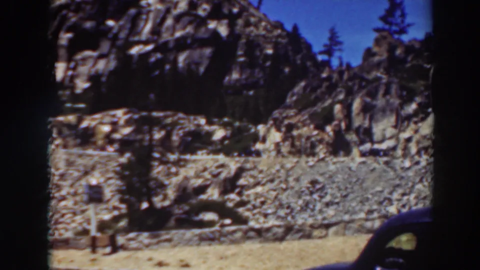 1937 Lake Tahoe frame showing a gravelly mountain roadside, rock face, pine trees, small roadside signs, and the cropped front edge of a dark automobile.