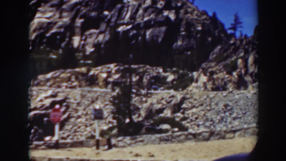 1937 Lake Tahoe home-movie frame showing small roadside signs, sunlit rocky terrain, pine trees, dry roadside ground, and the cropped edge of a dark vintage car.