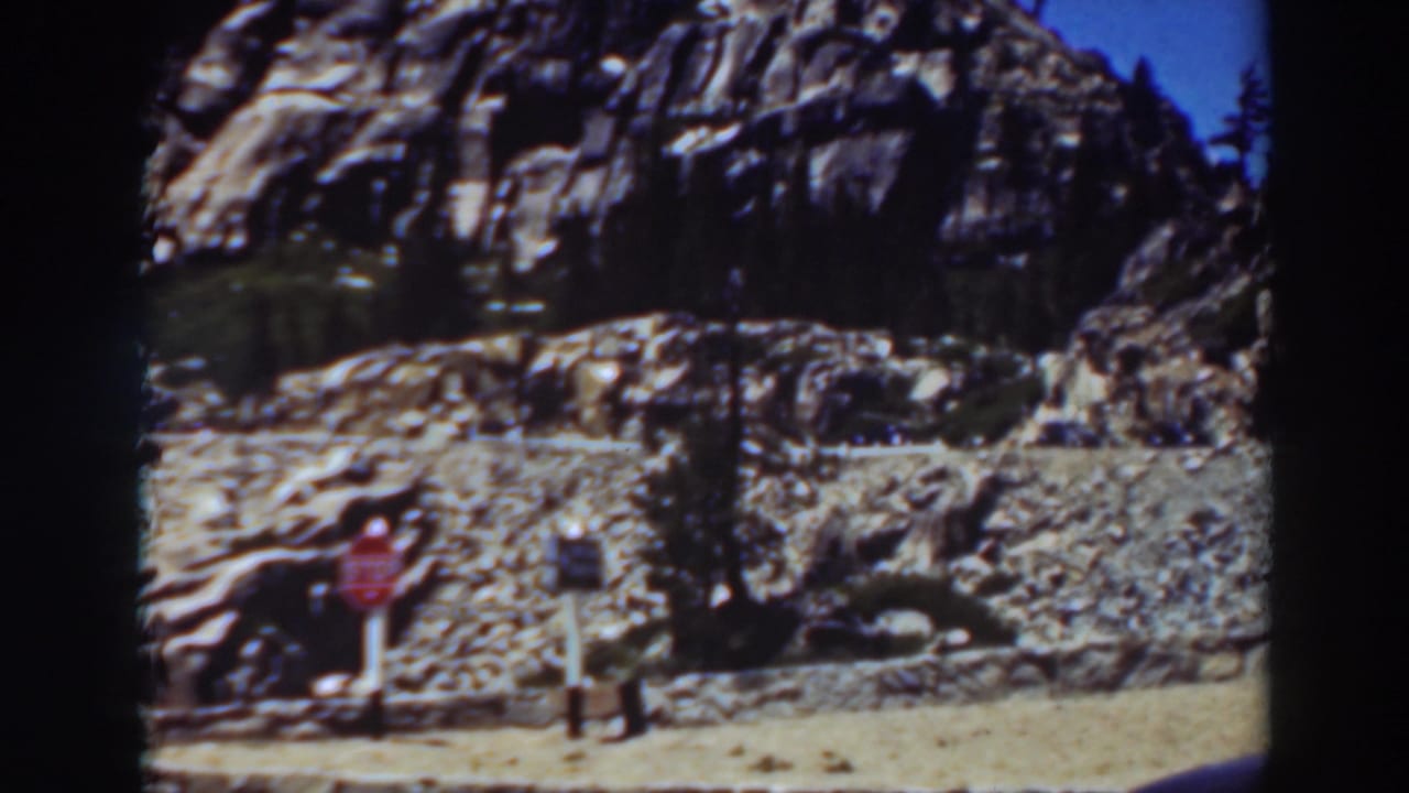 1937 Lake Tahoe-area roadside view with a stop sign at the base of a rocky slope and sparse trees in bright daylight.