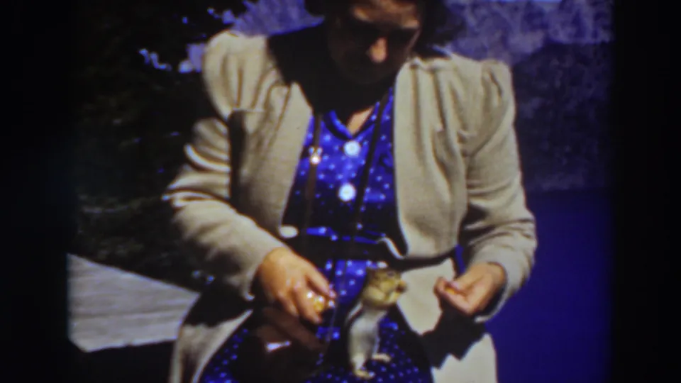 A seated woman at Lake Tahoe in 1937 looks down as a chipmunk remains on her lap near a small bag of food.