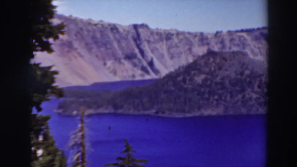 Lake Tahoe home-movie frame showing blue water, evergreen silhouettes, a wooded shoreline, and rocky mountain terrain under clear sky.