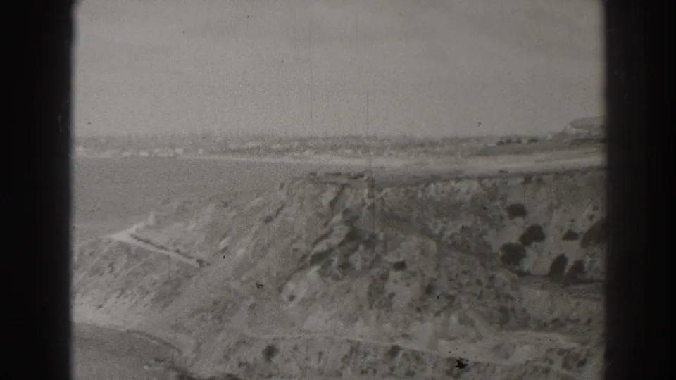 Black-and-white 8mm frame showing a Palos Verdes-area bluff, ocean water, and a narrow road or path across the slope.