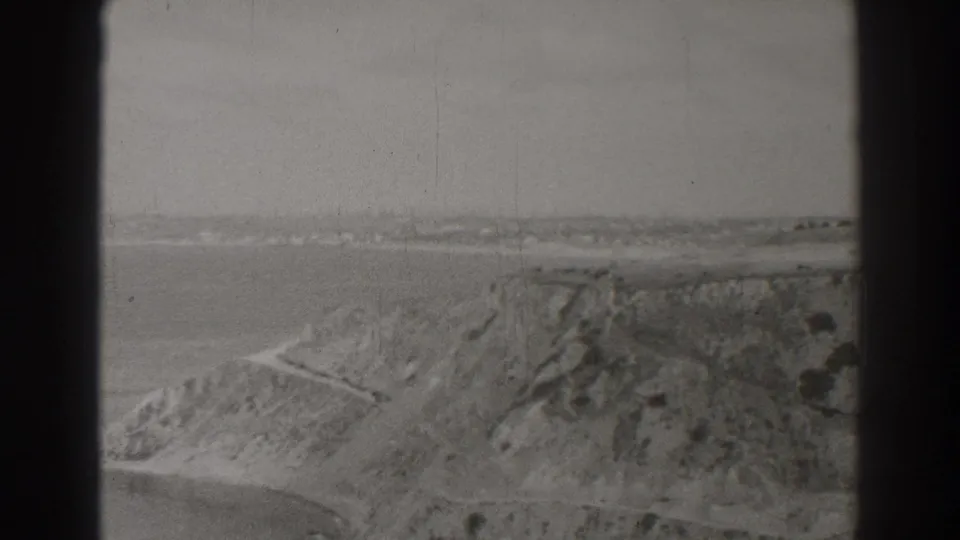 1937 Southern California coastal view with a rugged hillside, ocean water, and a light road line crossing the bluff.