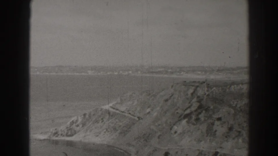 Black-and-white 8mm view of the Long Beach and Palos Verdes shoreline area, showing ocean, bluff terrain, and a winding road.