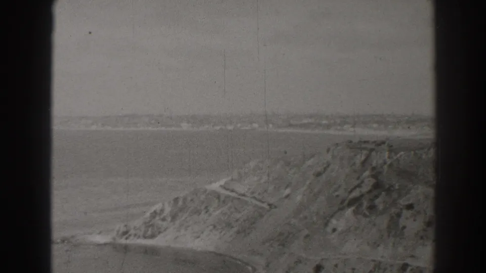 1937 Long Beach home-movie frame showing a rugged coastal bluff, cliffside path, calm water, and hazy distant shore.