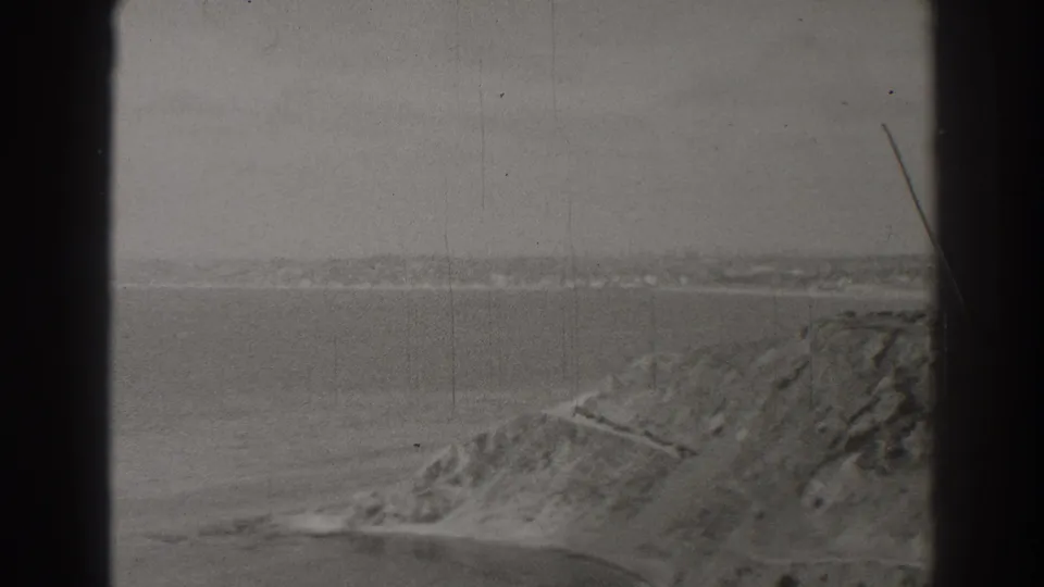 Black-and-white 8mm frame of the Long Beach coast, with a cliffside path, broad water, and distant shoreline.