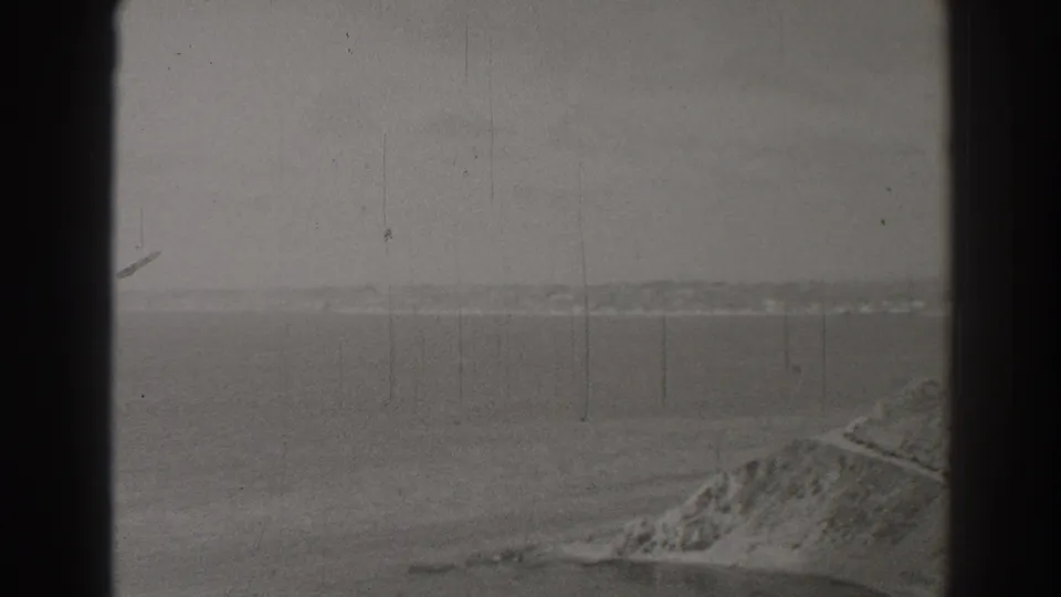 1937 Long Beach home-movie frame looking across calm water toward a hazy coast or mountain ridge, with rocky shoreline at lower right.