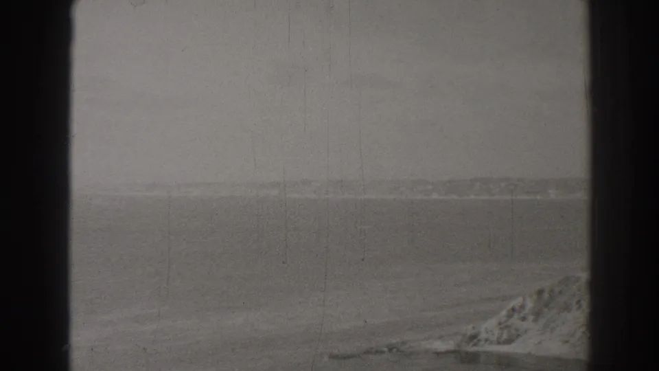 Black-and-white 8mm view from Long Beach across a hazy sea toward distant land, with a rocky shoreline edge visible in the foreground.