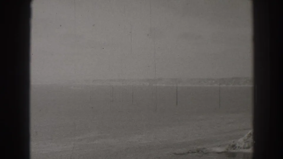 1937 8mm frame showing a muted Long Beach coastal horizon, broad water, and a cropped rocky shoreline under cloudy conditions.
