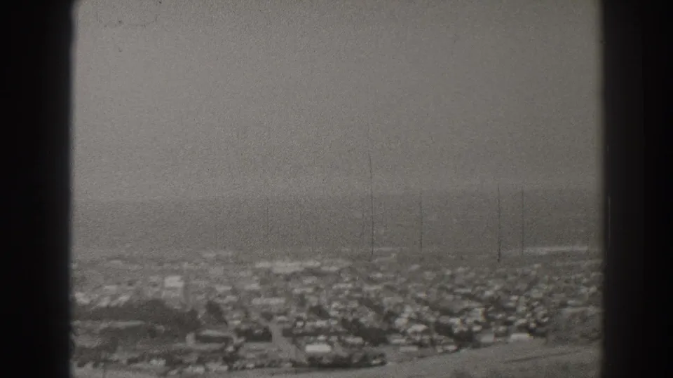 A 1937 Long Beach aerial-style frame shows compact housing, an open strip in the foreground, and a faint ocean line beyond.