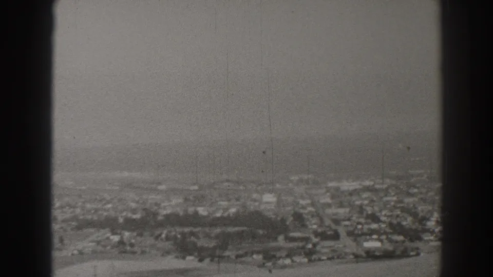Elevated black-and-white view of Long Beach with clustered houses, road grids, trees, haze, and visible 8mm frame edges.