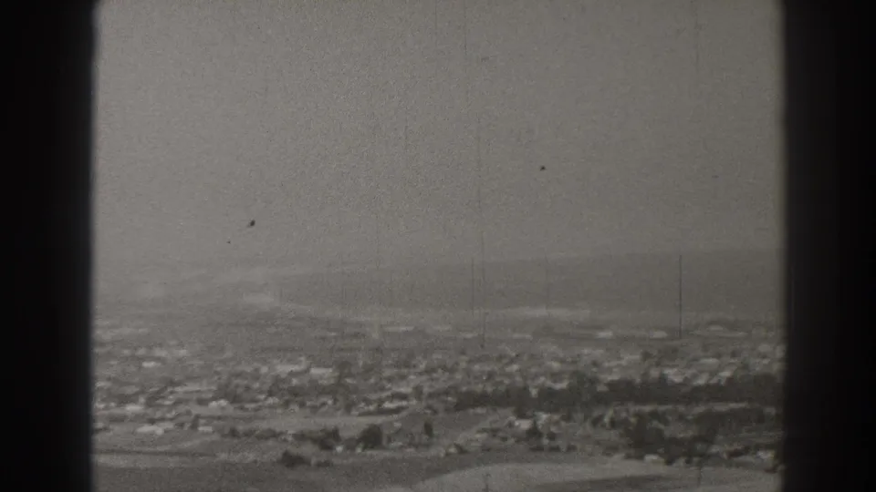 Elevated Long Beach view with rows of homes, pale open land, faint roads, and pronounced black-and-white 8mm grain.