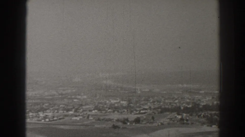 Black-and-white 8mm frame of Long Beach showing a hazy residential spread, road lines, trees, and vertical projector borders.