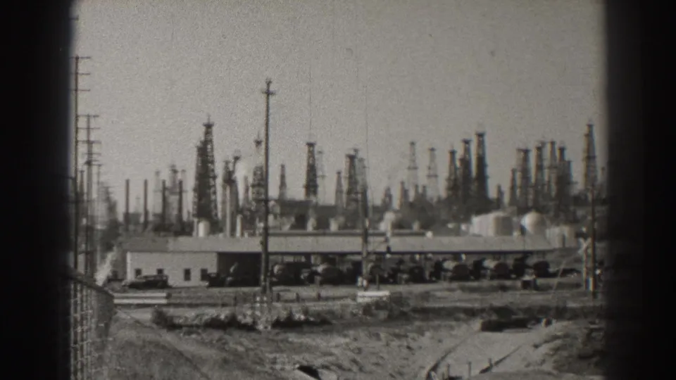 Wide 1937 Long Beach oil-field view with many derricks behind low industrial buildings, utility poles, and tanks or railcars.