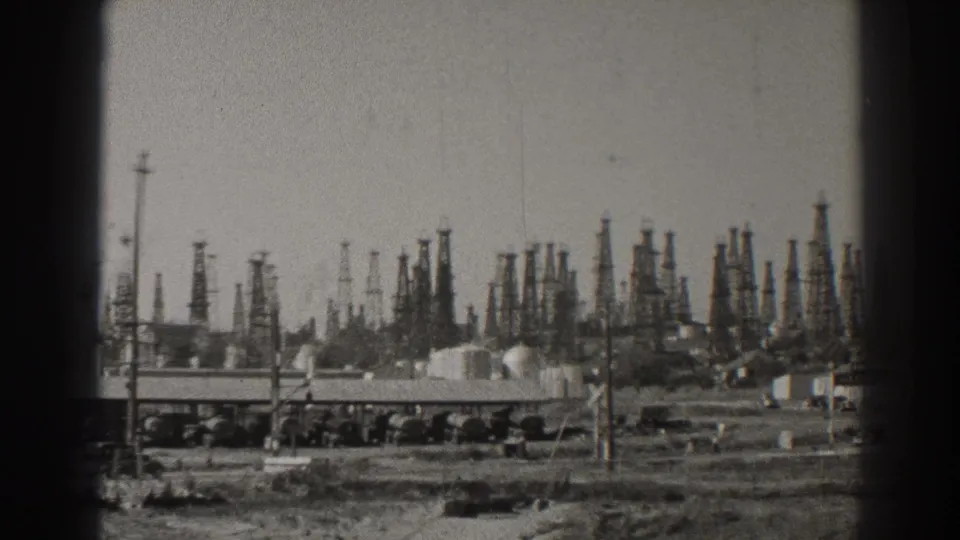 1937 Long Beach oil-field view with dense derricks, low industrial buildings, utility poles, and bare ground in the foreground.
