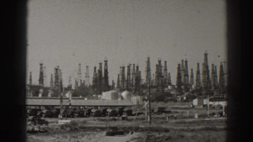 1937 Long Beach oilfield view with dense derricks beyond a roadside foreground, low buildings, tanks, and utility poles.