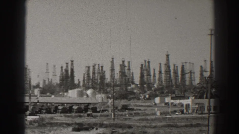 A 1937 Long Beach oilfield panorama shows derricks rising behind low industrial buildings, tanks, utility poles, and open lots.