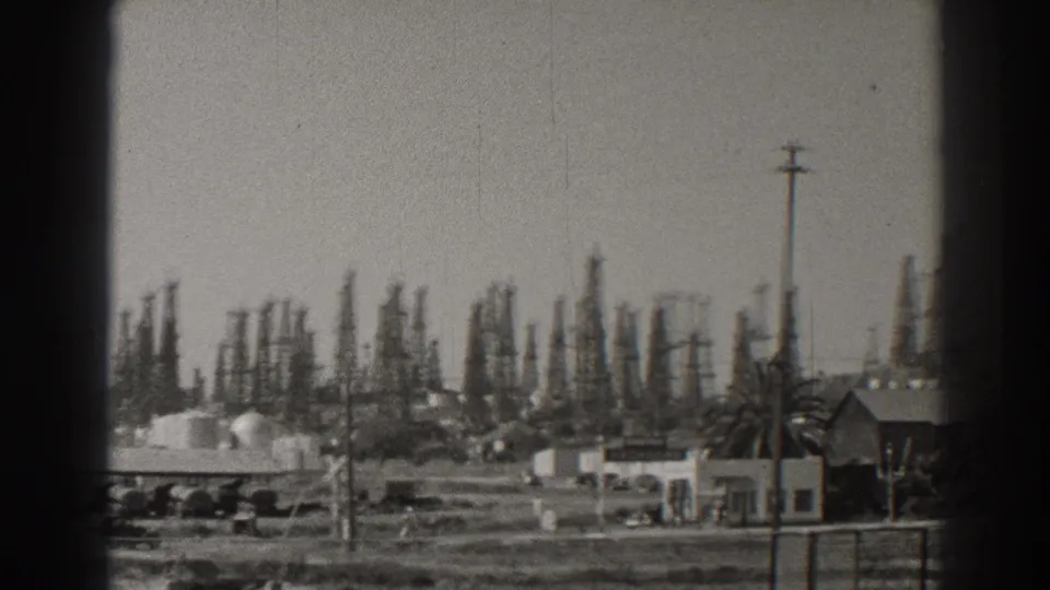 Long Beach oilfield footage shows a low roadside building and palm tree in front of dense derricks and utility poles.