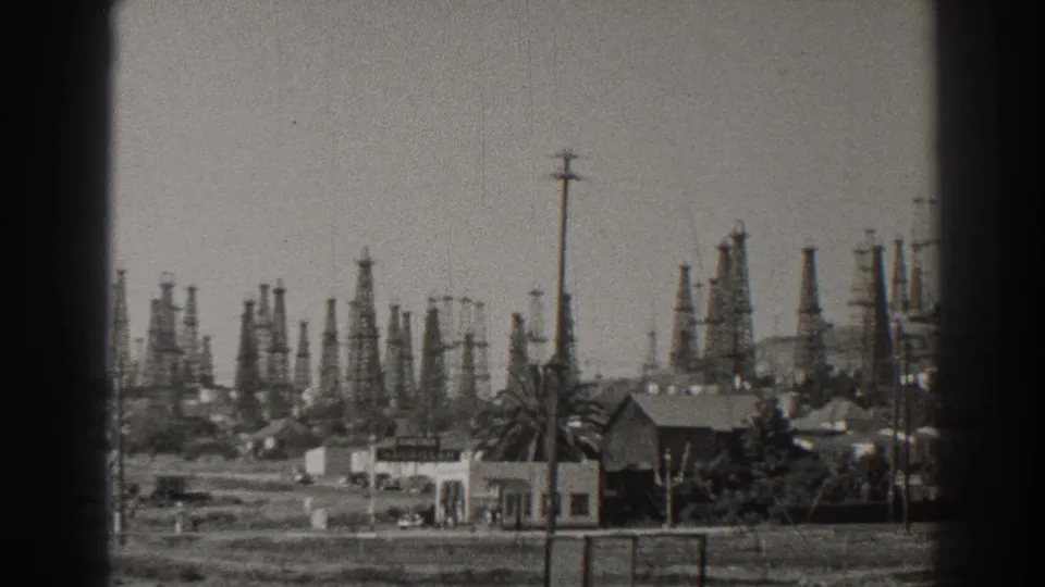 The 1937 Long Beach view frames oil derricks behind a low building, palm tree, fence line, utility poles, and open ground.