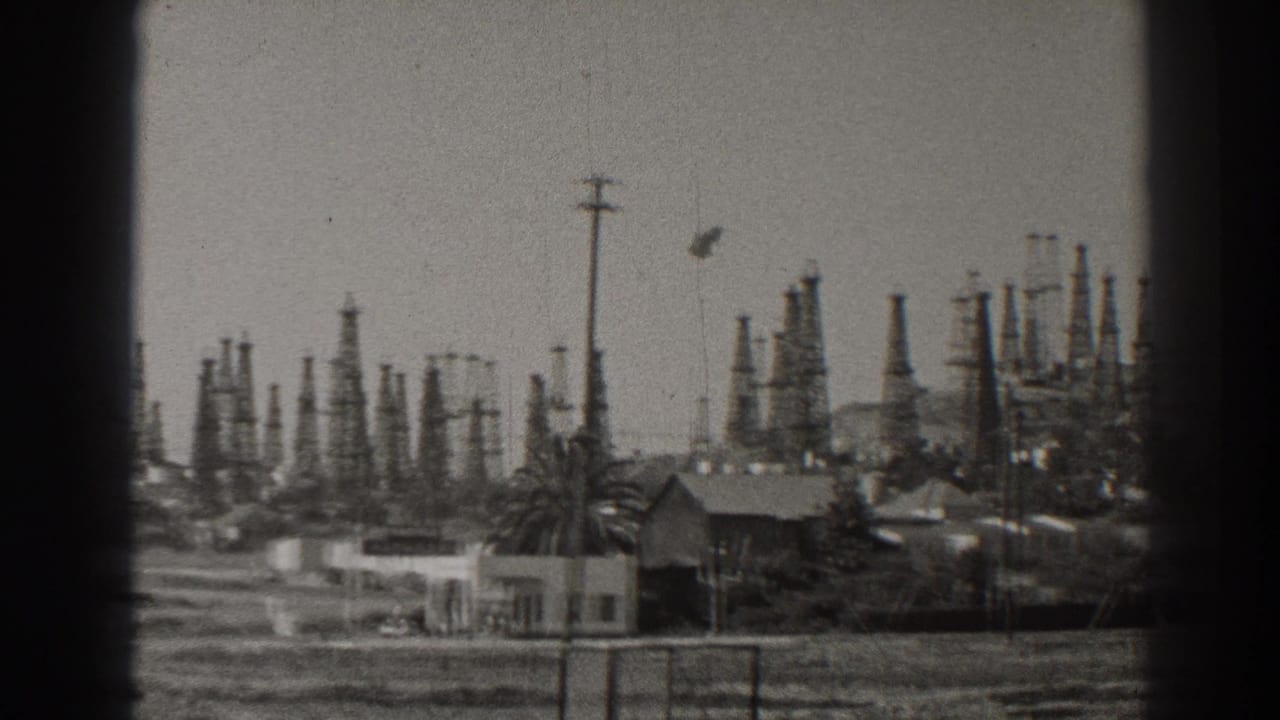 1937 Long Beach view with clustered oil rigs rising behind small buildings, palms, open ground, and utility poles.