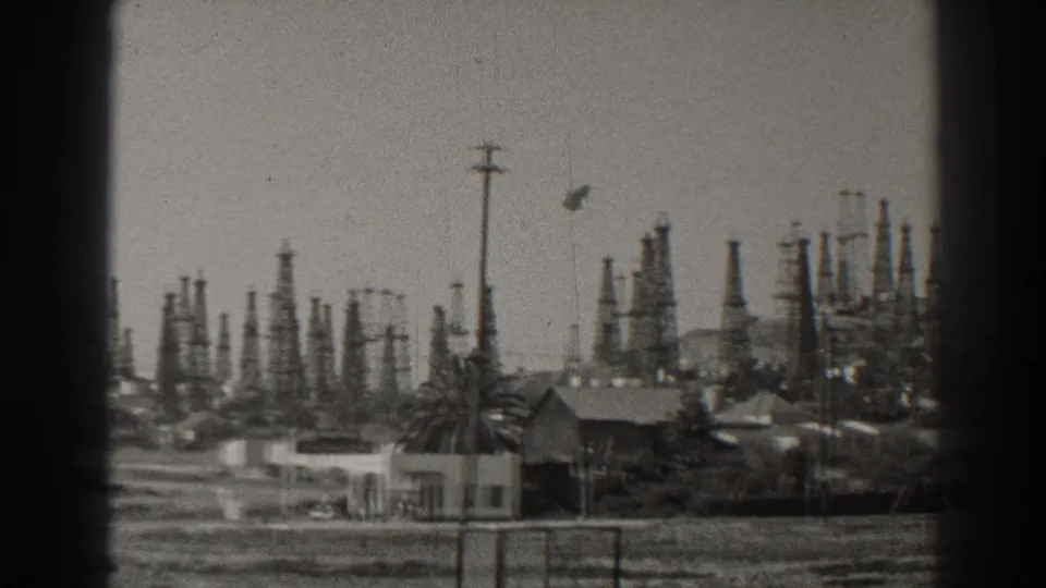 1937 Long Beach view with clustered oil rigs rising behind small buildings, palms, open ground, and utility poles.