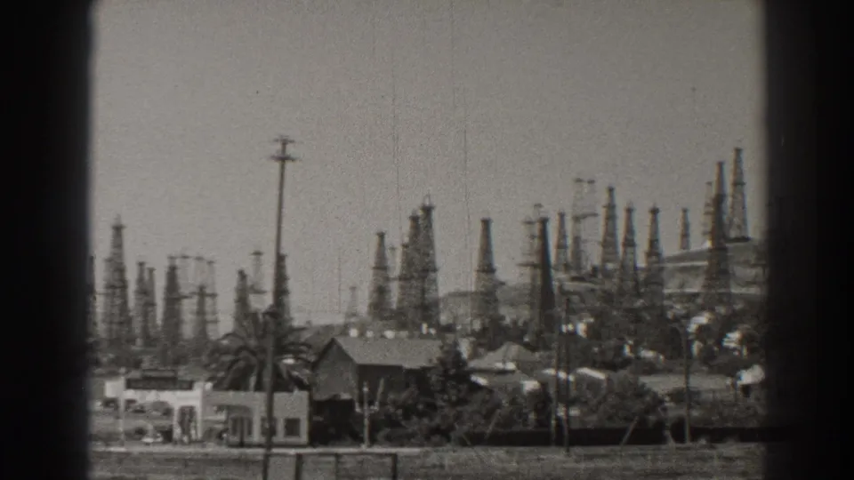 A Long Beach oil-field view shows derricks across the horizon with roadside buildings, palms, and poles in the foreground.