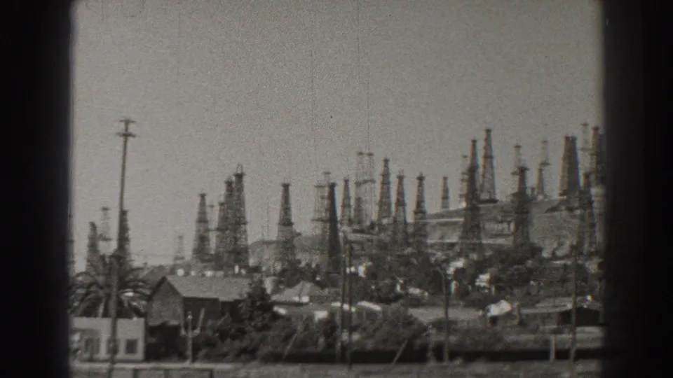 1937 Long Beach oil-field panorama with derricks rising behind rooftops, palms, utility poles, and brushy foreground.