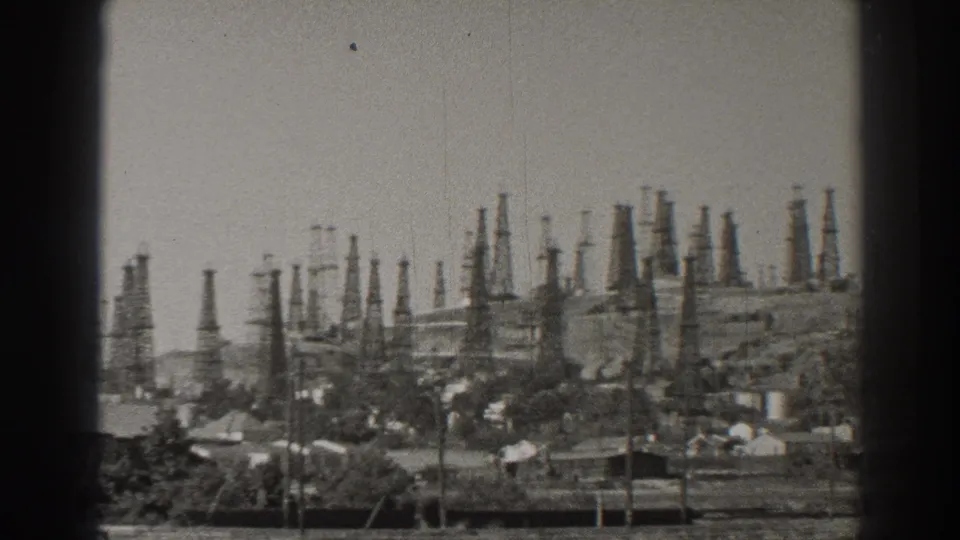Long Beach landscape showing numerous oil derricks among low structures, trees, utility poles, and distant raised ground.