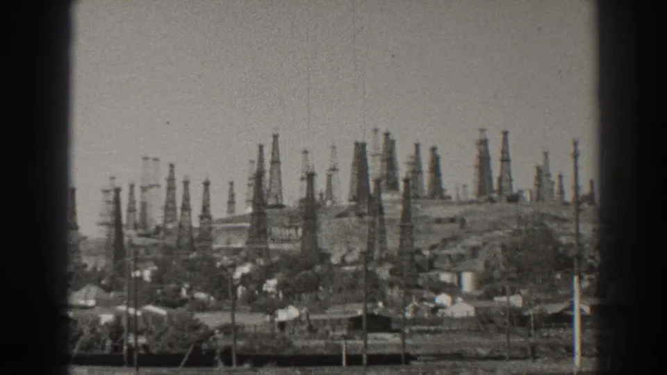 1937 Long Beach home-movie frame showing many oil derricks rising over a small neighborhood and utility poles.