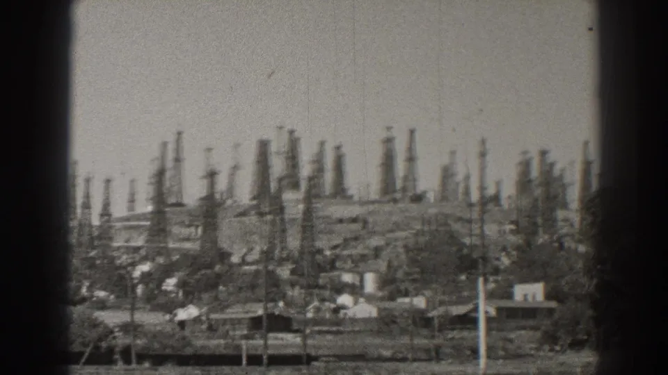 Long Beach, California frame with crowded oil derricks on the slope behind houses and scattered trees.