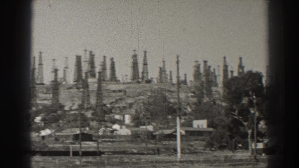 1937 Long Beach view of an oil-field hillside, with derricks behind houses, poles, and a prominent tree.