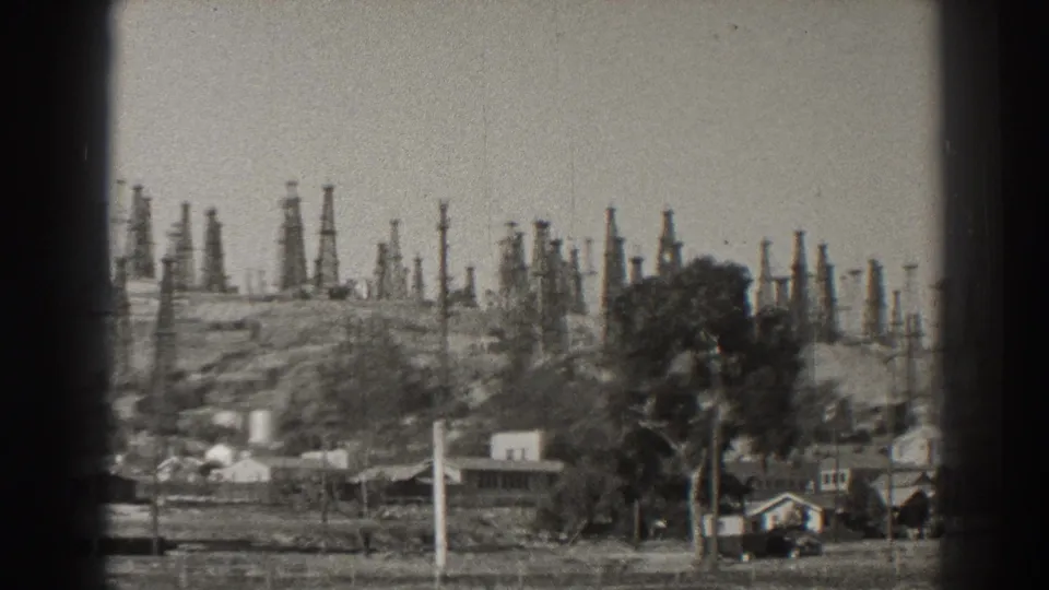Long Beach oil derricks stand across the hillside behind trees, poles, and a small neighborhood building.