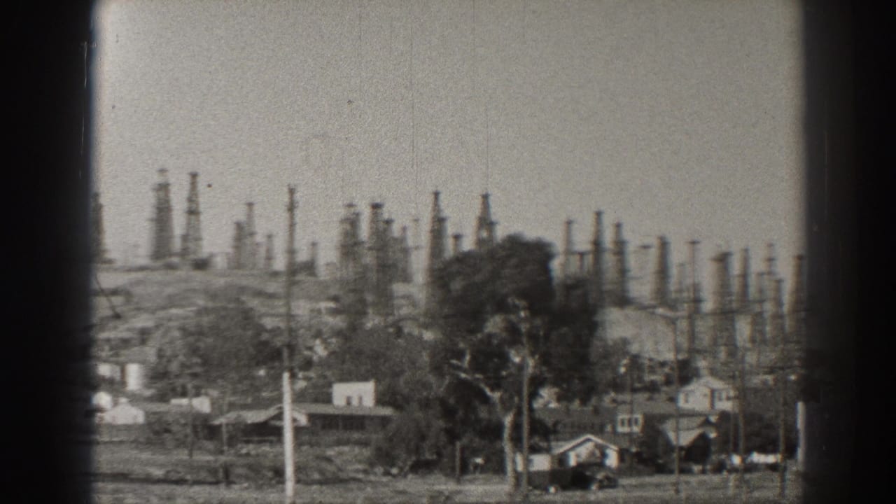 Distant oil derricks behind houses and utility poles on a hillside.