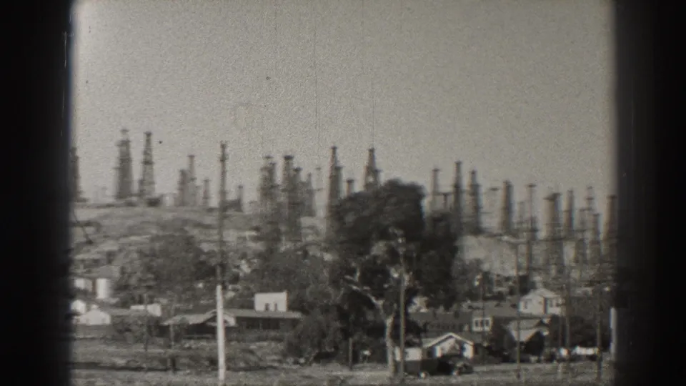1937 Long Beach view with oil derricks rising behind small houses, utility poles, and hillside terrain in black-and-white 8mm footage.