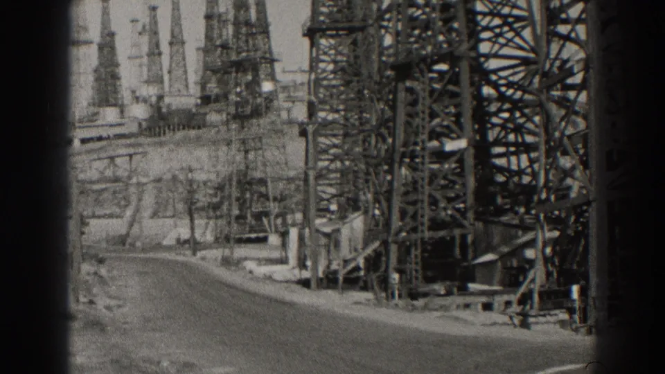 Black-and-white 1937 Long Beach frame showing a paved road curving past tall lattice towers and oil-field equipment.