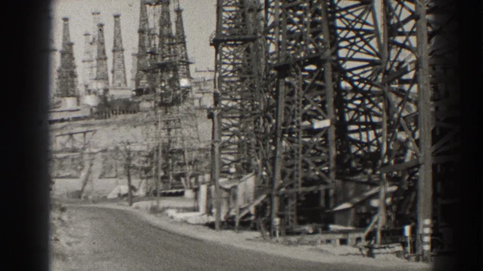 1937 Long Beach oil-field scene with dense steel framework, derricks, and a paved road in soft black-and-white 8mm texture.