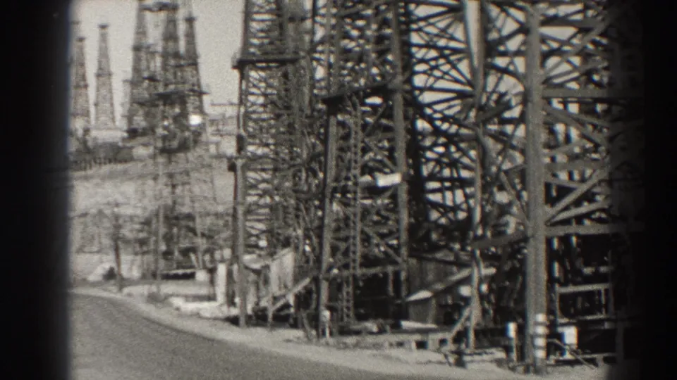 Black-and-white 1937 Long Beach frame with close oil-field lattice structures, distant derricks, and the edge of a paved road.