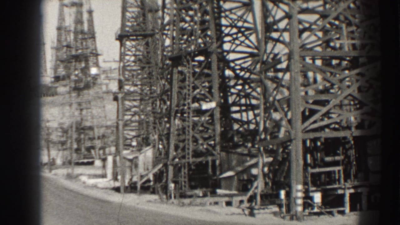 Black-and-white view of steel oil derricks beside a road or yard edge.