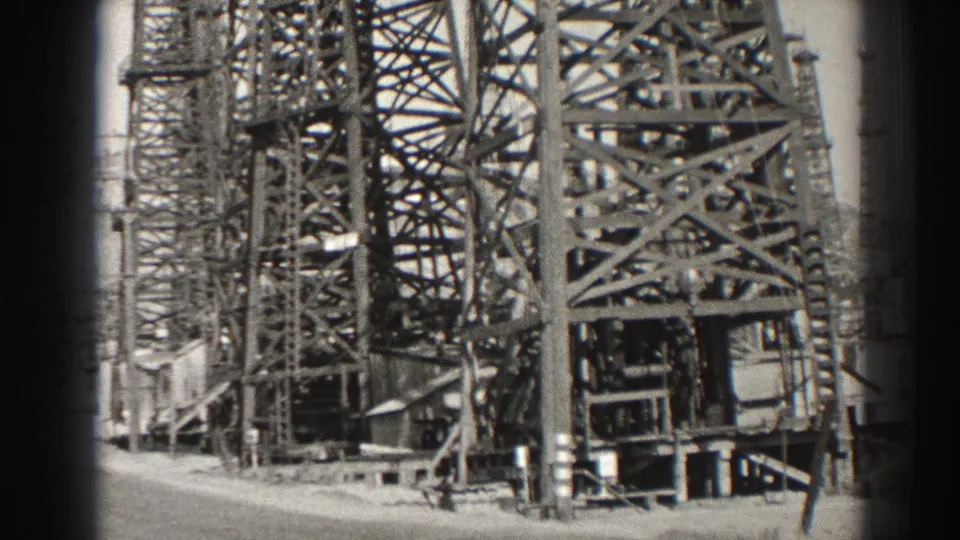 1937 Long Beach oil-field frame showing closer steel derrick framing, lower supports, and equipment near a road edge.