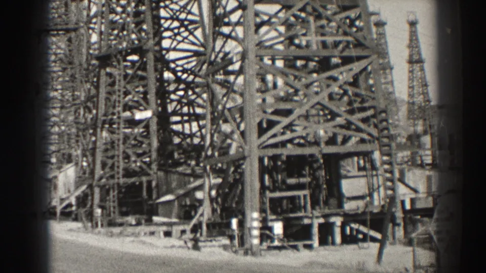 Black-and-white 8mm frame from Long Beach in 1937 showing oil derricks, pump structures, and bright exposed ground.