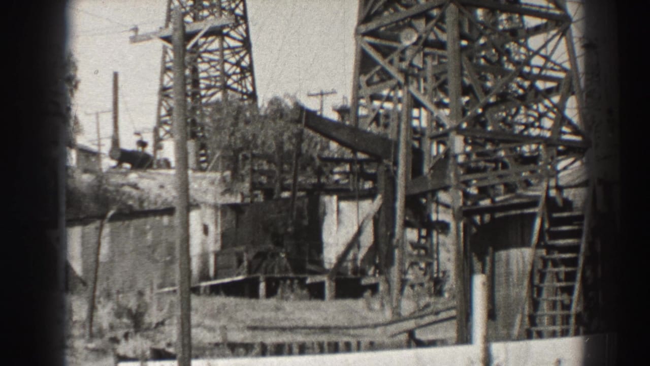 Black-and-white view of an oil derrick with ladder, platform, utility poles, and a small base structure.